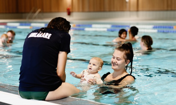 Simlärare sitter på bassängkanten, mamma med barn är precis nedanför i vattnet.