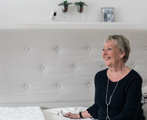 Äldre kvinna sitter i soffan i ett ljust rum. På hyllan ovanför henne står en vit, diskret tillsynskamera.