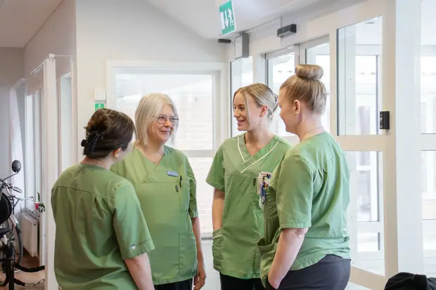 Fyra kvinnor i gröna vård-tunikor står i en ljus korridor och pratar.